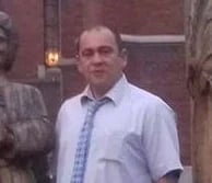 Man wearing glasses and light blue shirt standing outdoors against a brick wall background