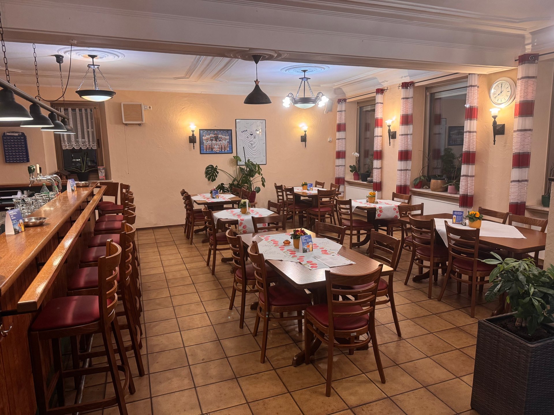 Innenraum eines gemütlichen Restaurants mit Holztischen, Bar-Sitzplätzen, warmem Licht und Topfpflanzen